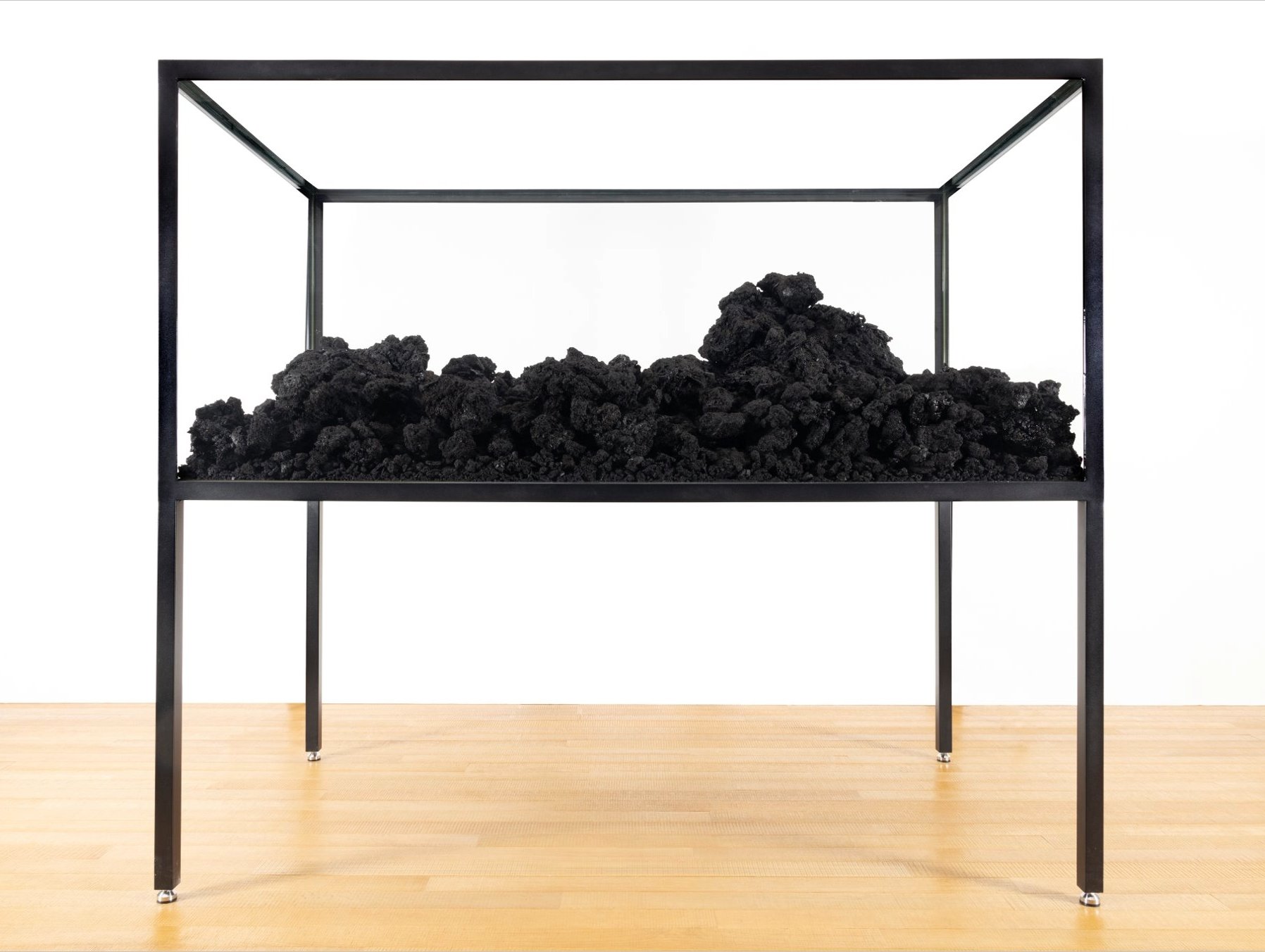 Clods of irregular-shaped black residue are displayed in a rectangular, black-framed glass case supported by four thin legs. The black residue fills approximately a quarter of the case and resembles soil.